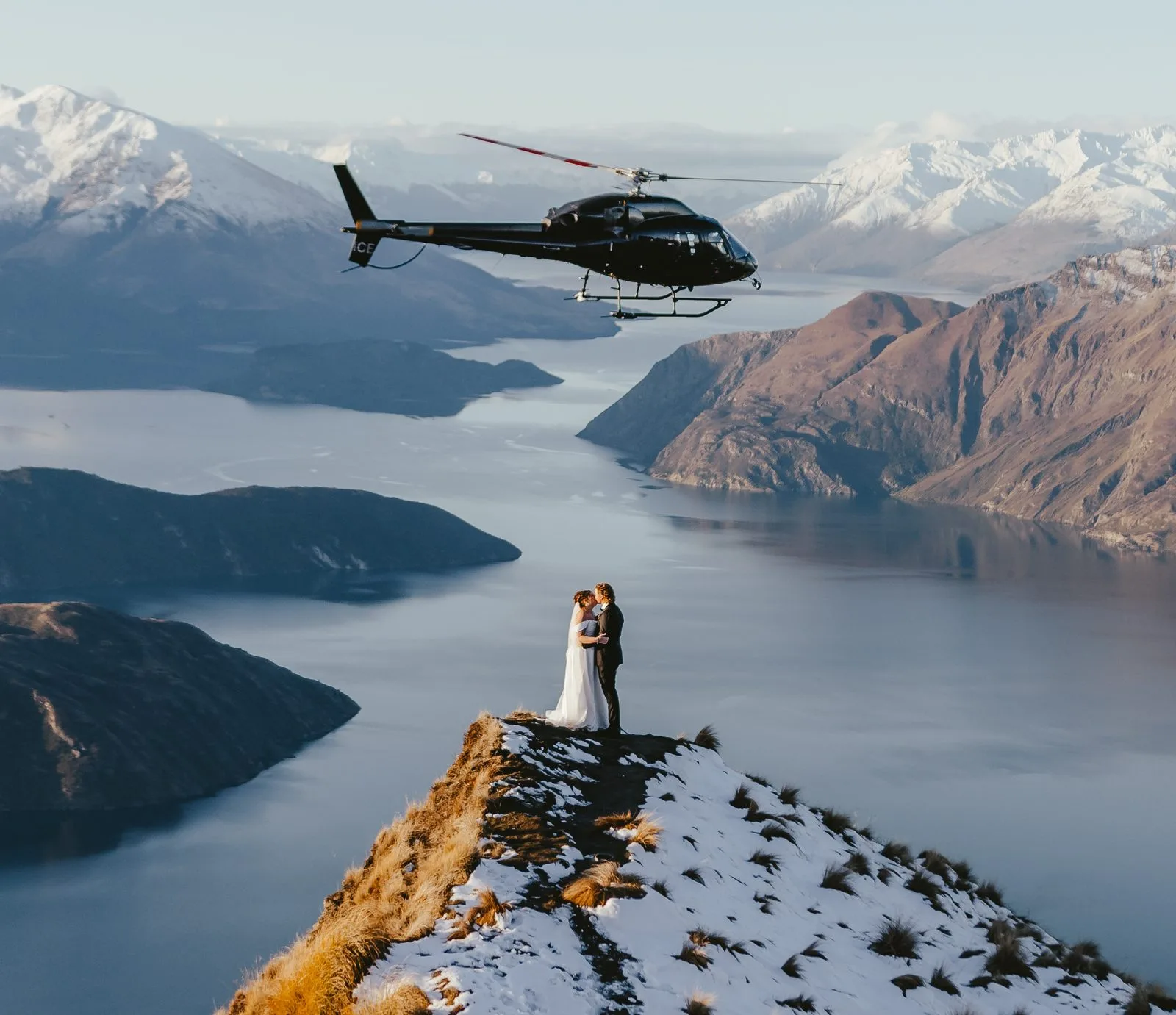 Couple on the snow-dusted Coromandel Peak ridge with a helicopter overhead and Lake Wānaka below