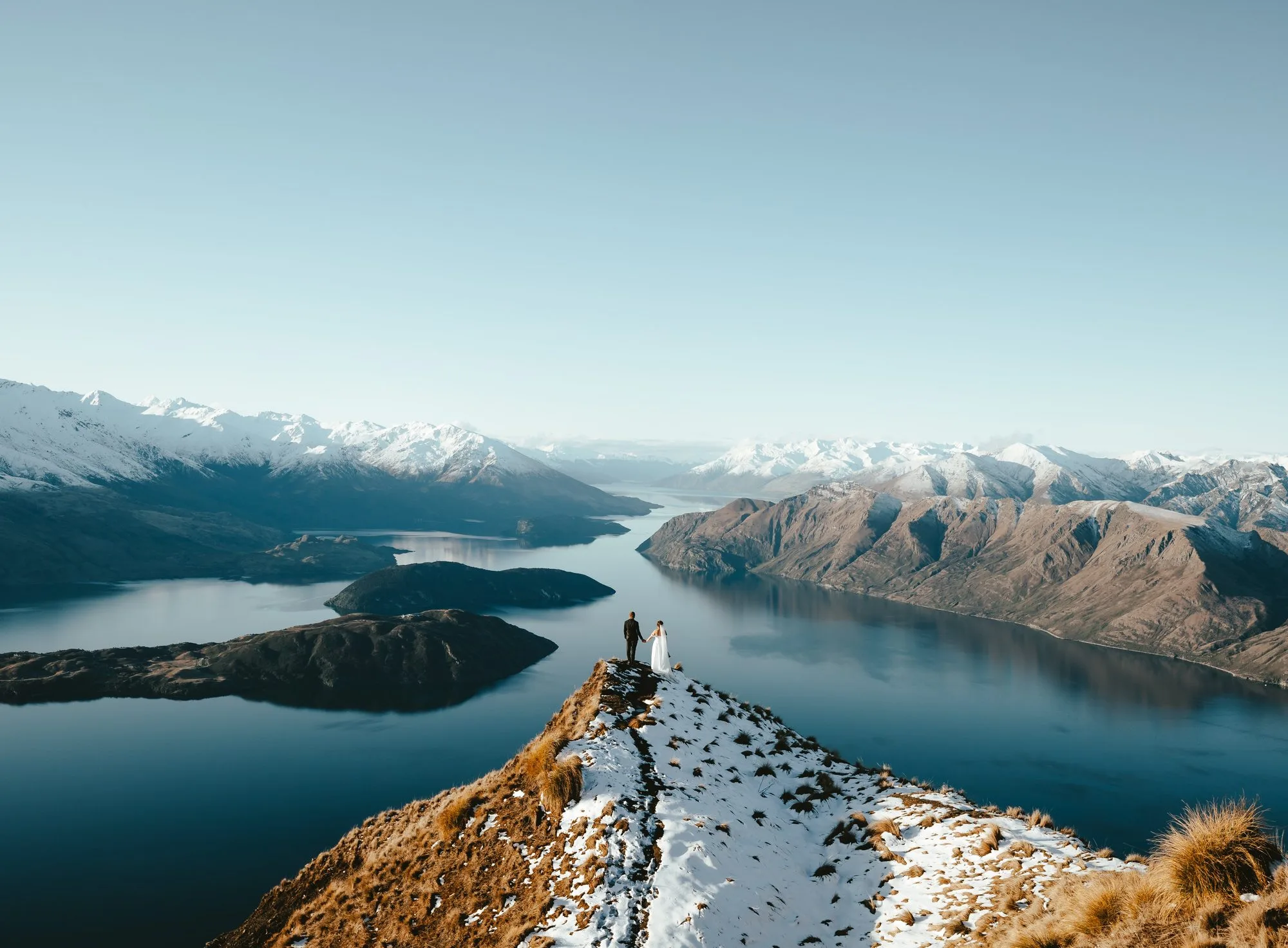 Couple standing hand in hand on the Coromandel Peak ridgeline high above Lake Wānaka, snow on the surrounding mountains