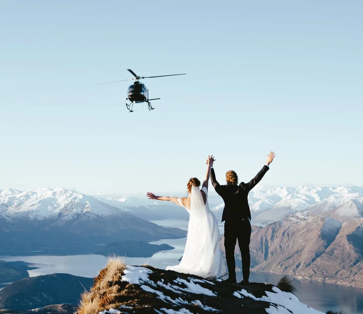 Couple with arms raised on Coromandel Peak as a helicopter flies over Lake Wānaka